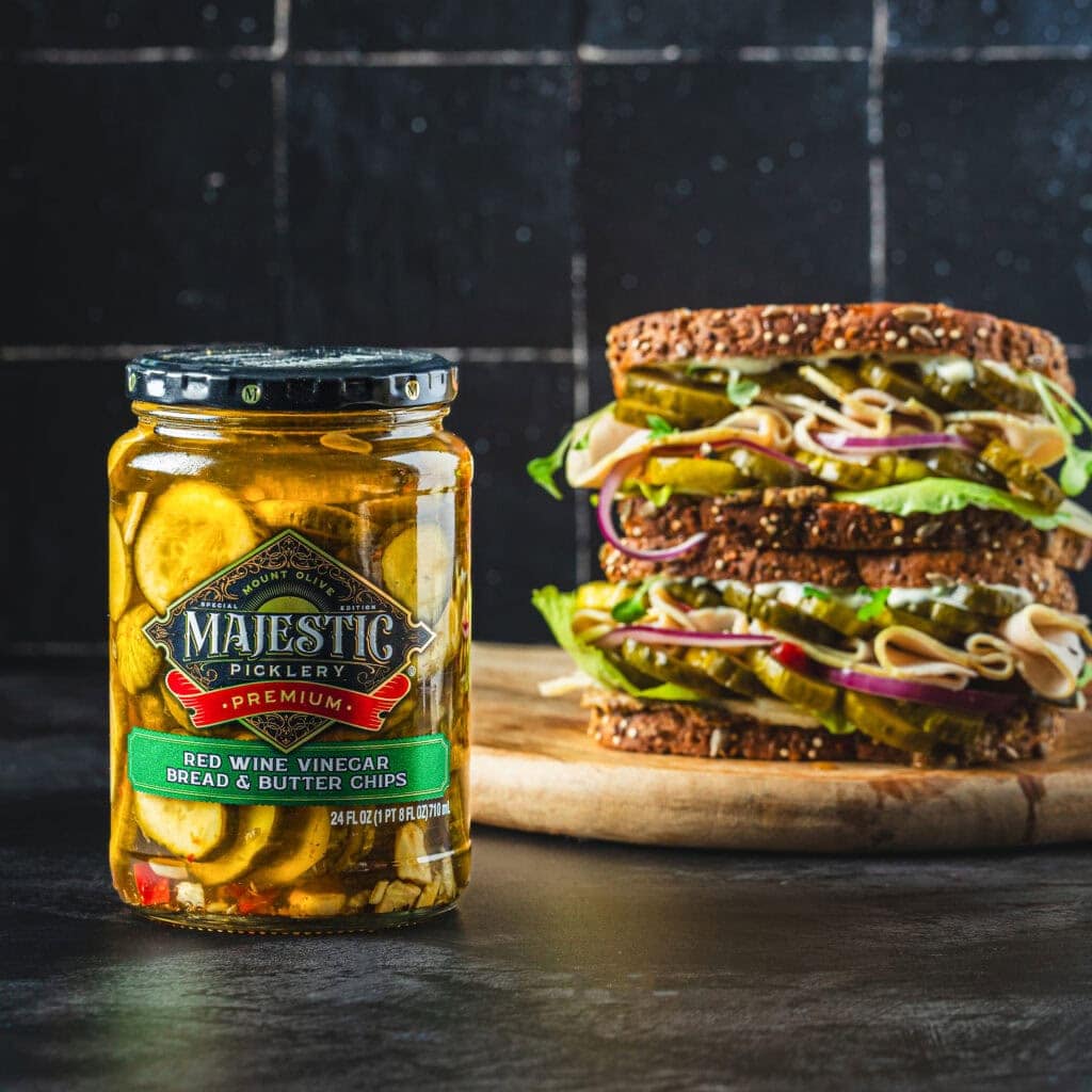 jar of Majestic bread and butter chip pickles with sandwich on a wood cutting board in the background