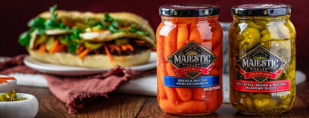 Assorted Majestic Picklery pickles and a sandwich on a rustic table.