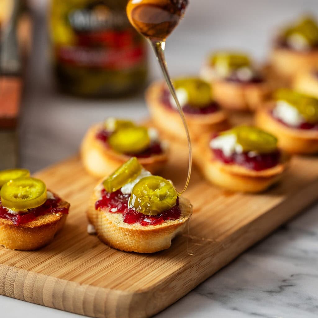 close up of cutting board showing mini crostini peices covered in cheese, jam and honey topped with jalapeno slices