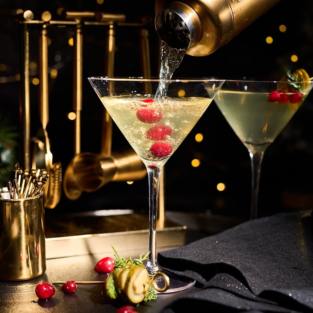 black background with two martini glasses. Martini is being poured from a gold cocktail shaker. garnished with berries and pickles