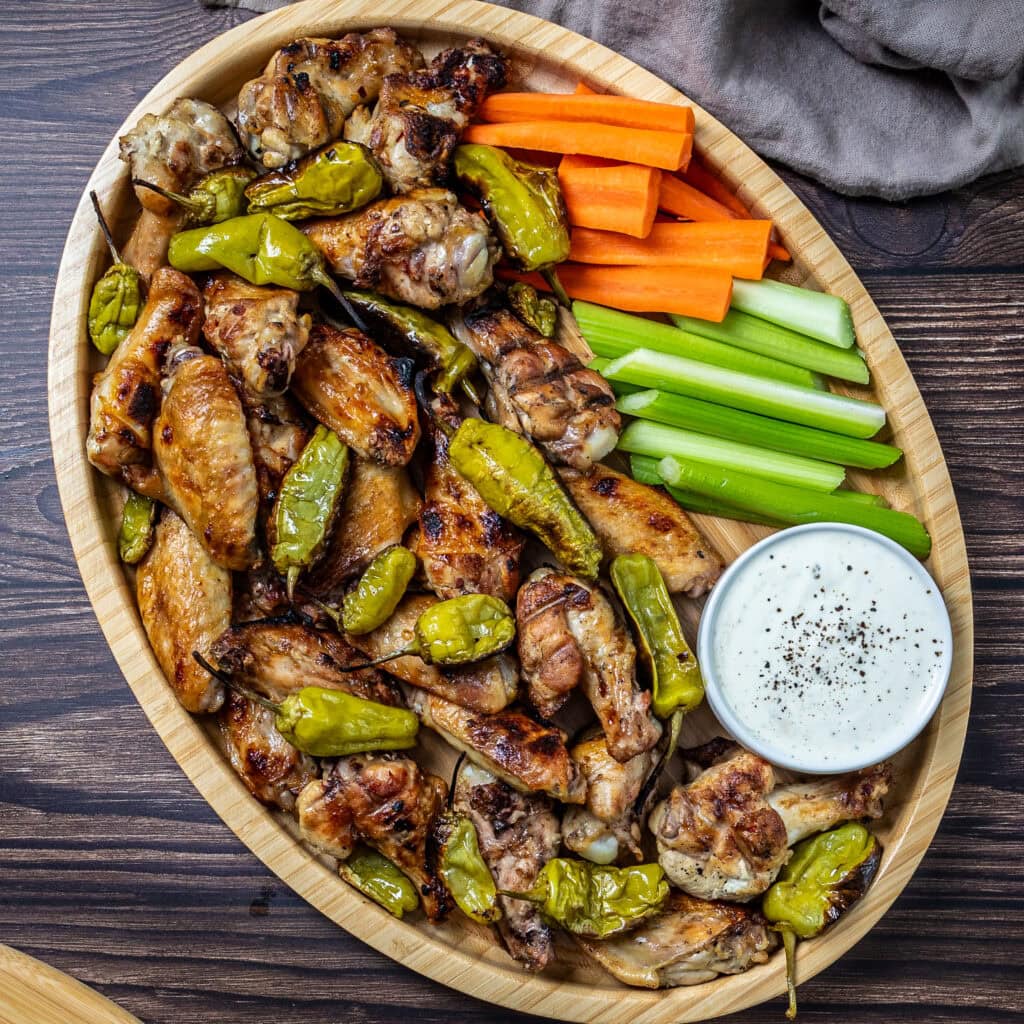 A wooden tray filled with grilled chicken wings, pepperoncini peppers, dish of ranch dressing, carrot sticks and celery sticks.