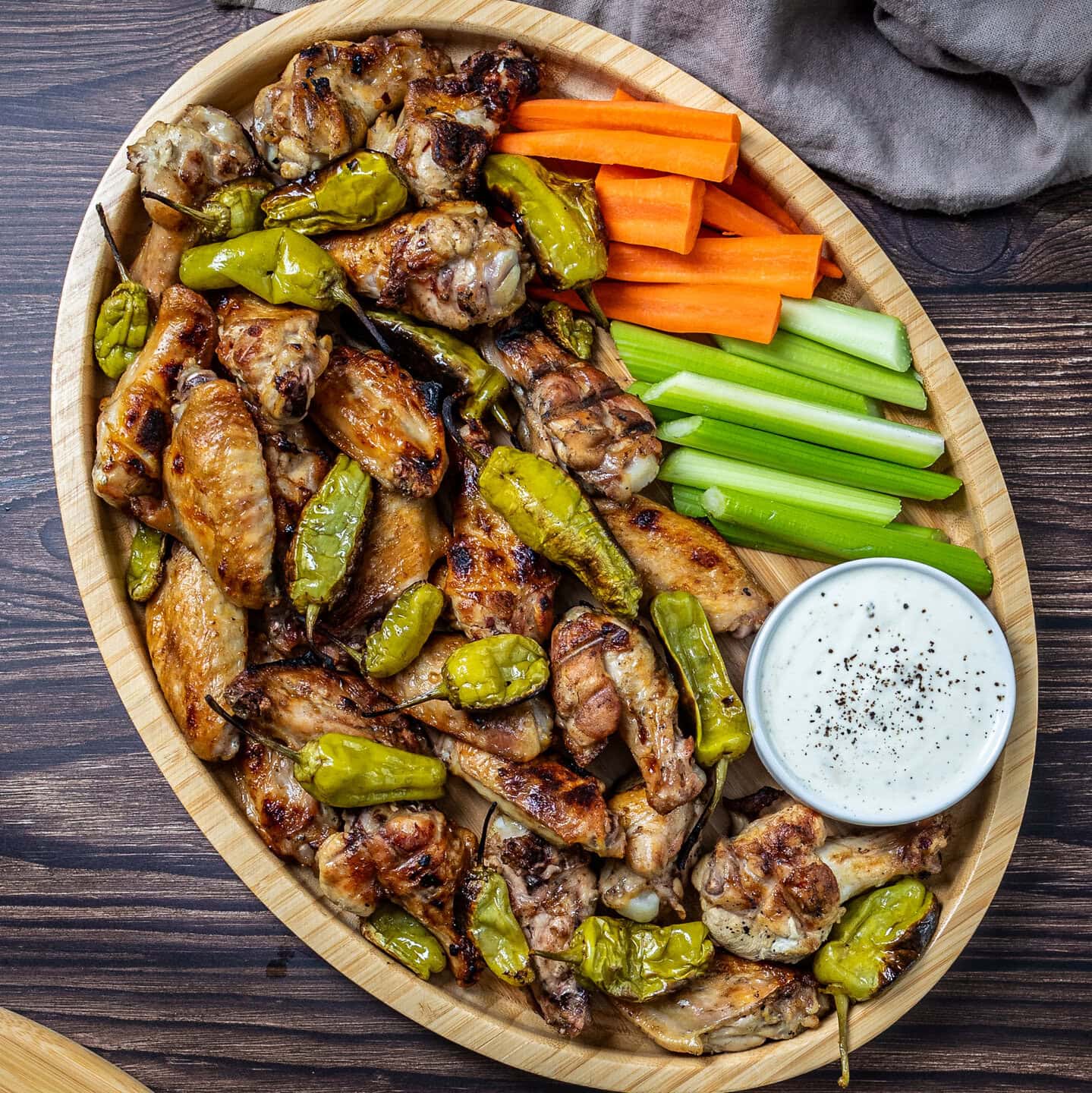 A wooden tray filled with grilled chicken wings, pepperoncini peppers, dish of ranch dressing, carrot sticks and celery sticks.