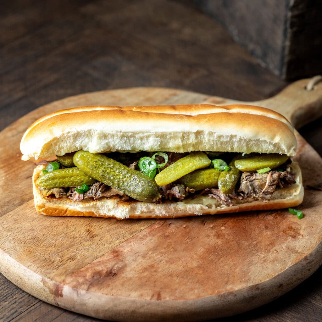 Dill Pickle Pot Roast on a hoagie roll topped with small pickles and sitting on a board.