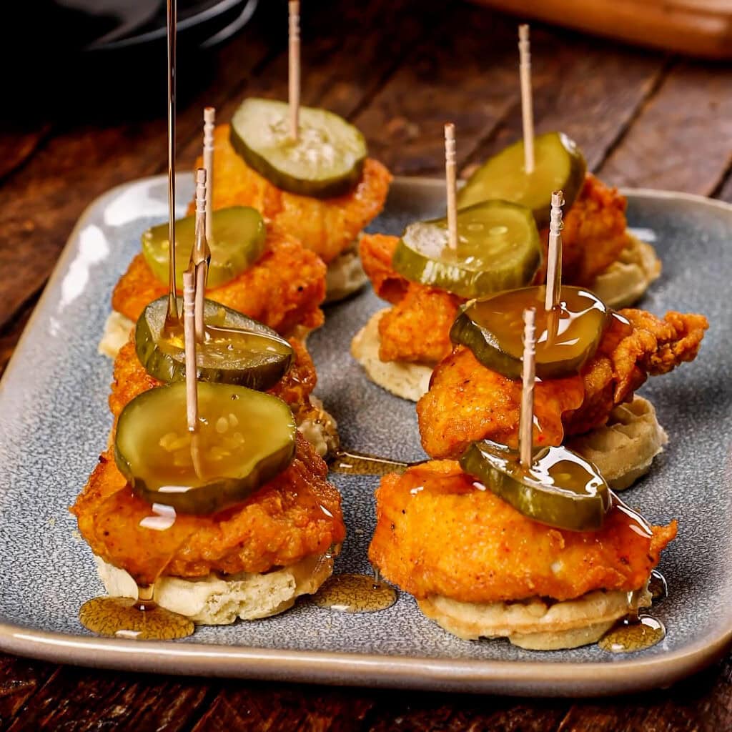 A plate showing mini waffles topped with chicken nuggets and pickle chips and a toothpick down the middle of each.