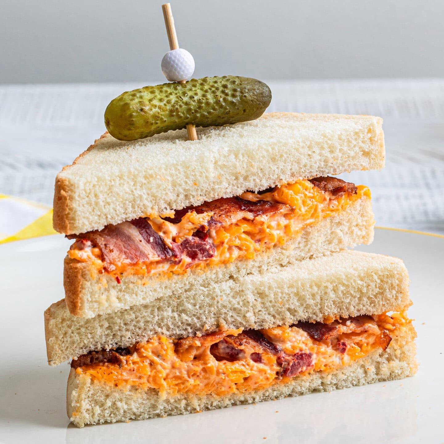 A sandwich with pimento cheese and bacon topped with a baby dill pickle on a white plate.