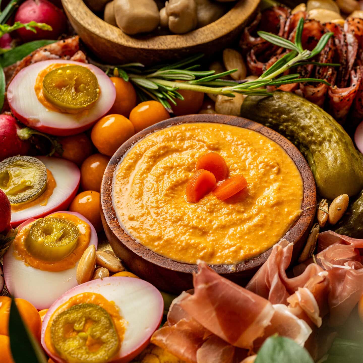 A charcuterie board close up, showing deviled eggs and a wooden dish filled with orange carrot hummus.