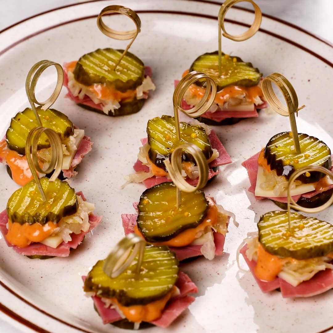 A white plate containing many Bread & Butter Reuben Sliders - little pickle sandwiches with toothpicks.