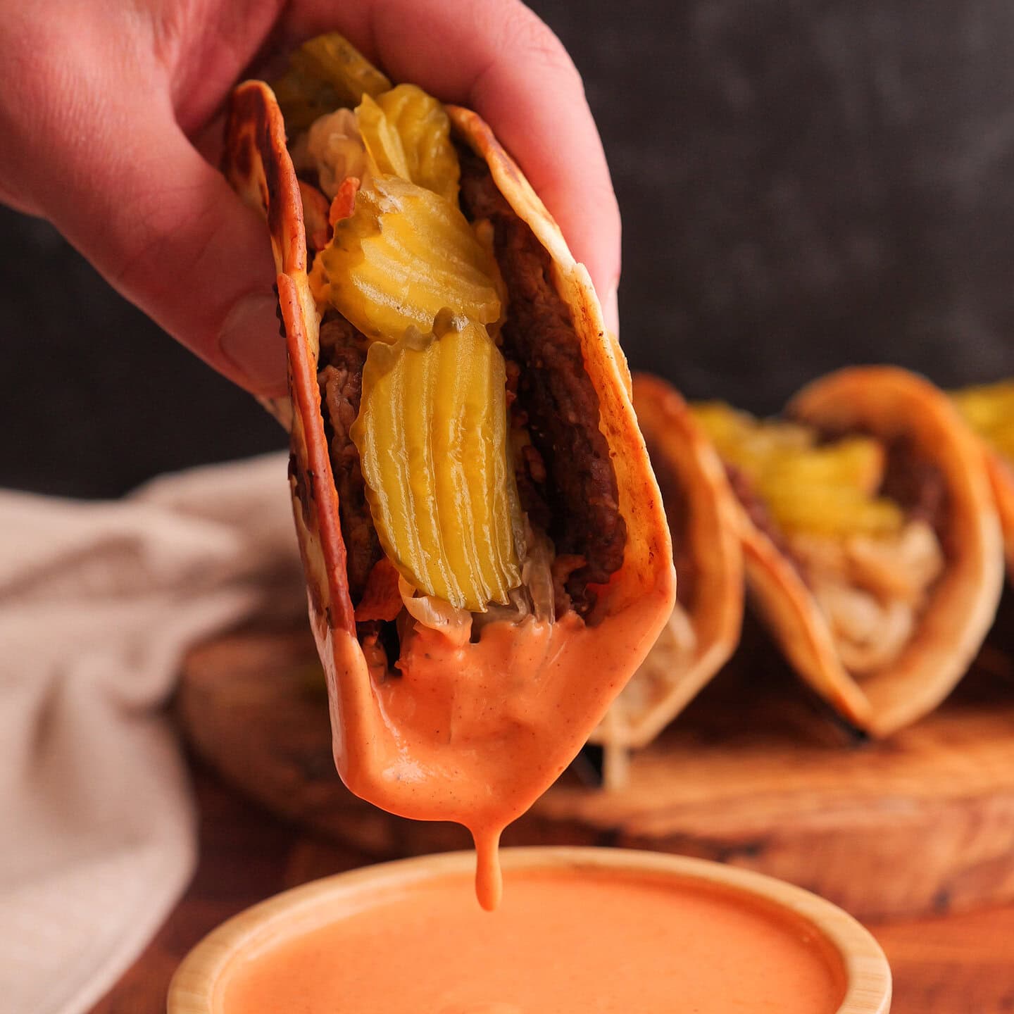 Smash burger taco topped with pickles being dipped into roasted red pepper sauce
