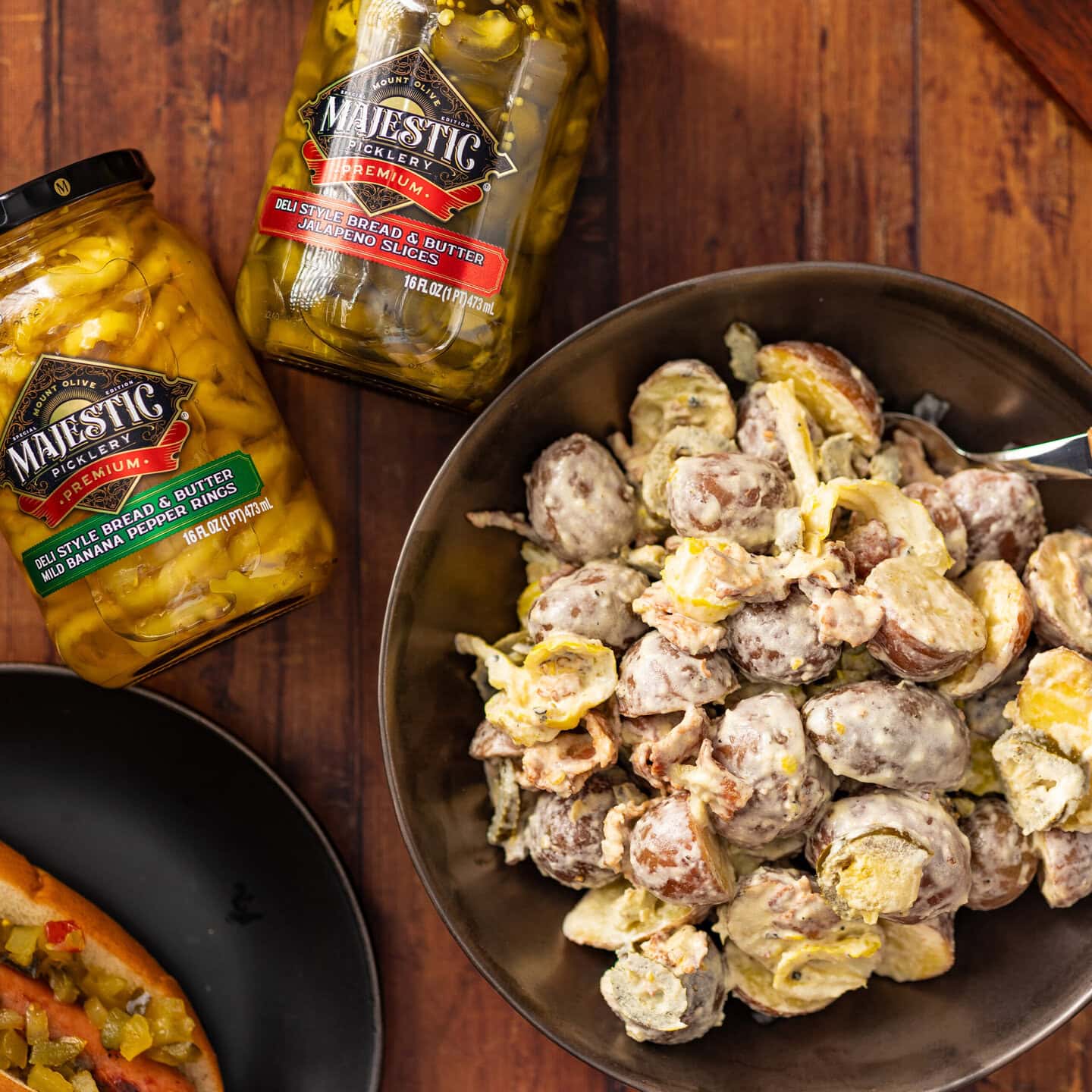 A bowl of potato salad next to two jars of Majestic Picklery jars on a wooden table.