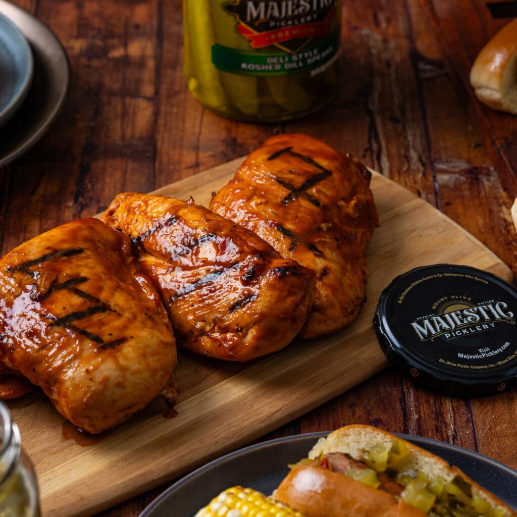 Three grilled chicken breasts on a wooden cutting board next to a Majestic Picklery jar lid.