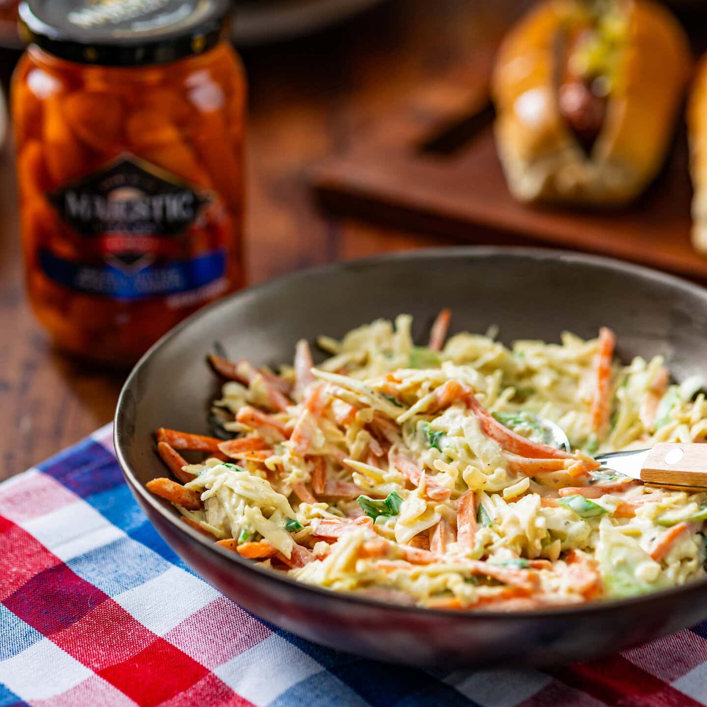 A bowl of coleslaw made with pickled carrots sitting on a red white and blue tablecloth on a wood table with jar of pickled carrots nearby.