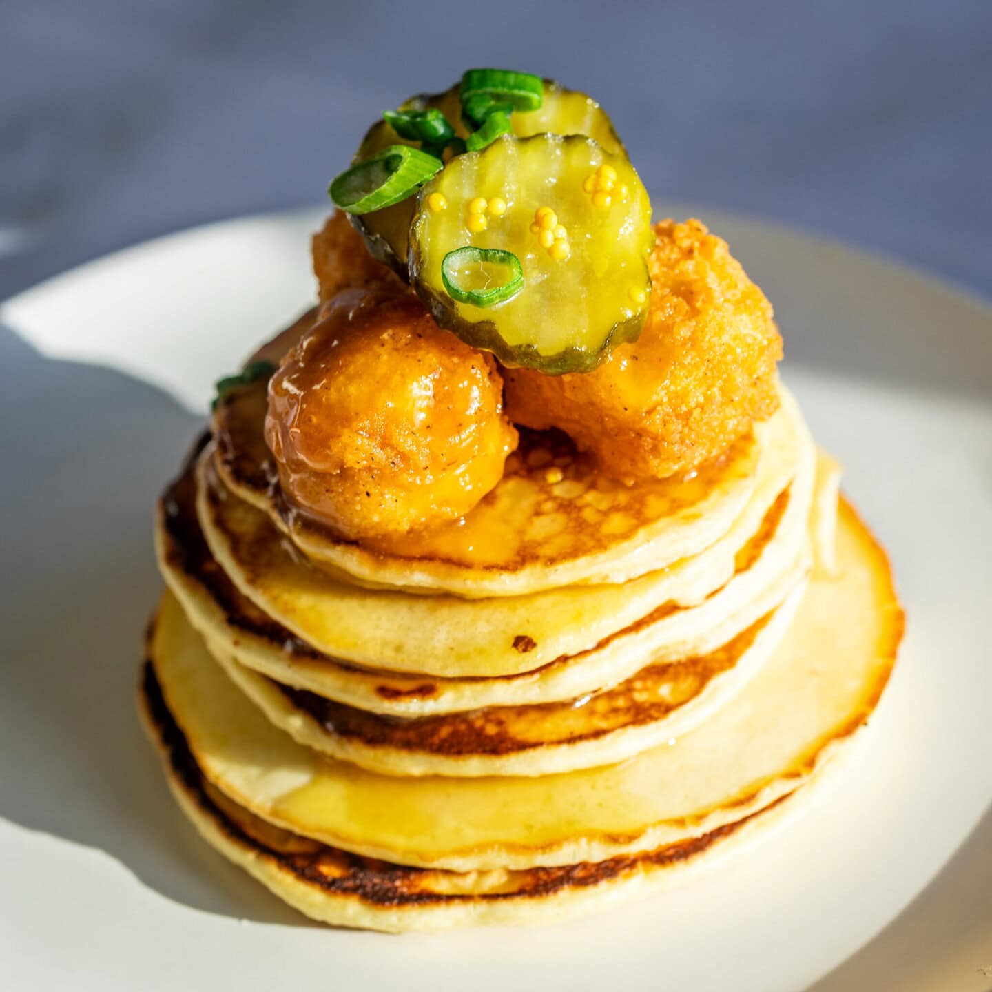 A stack of pancakes on white plate topped with popcorn chicken and pickles topped with hot honey.