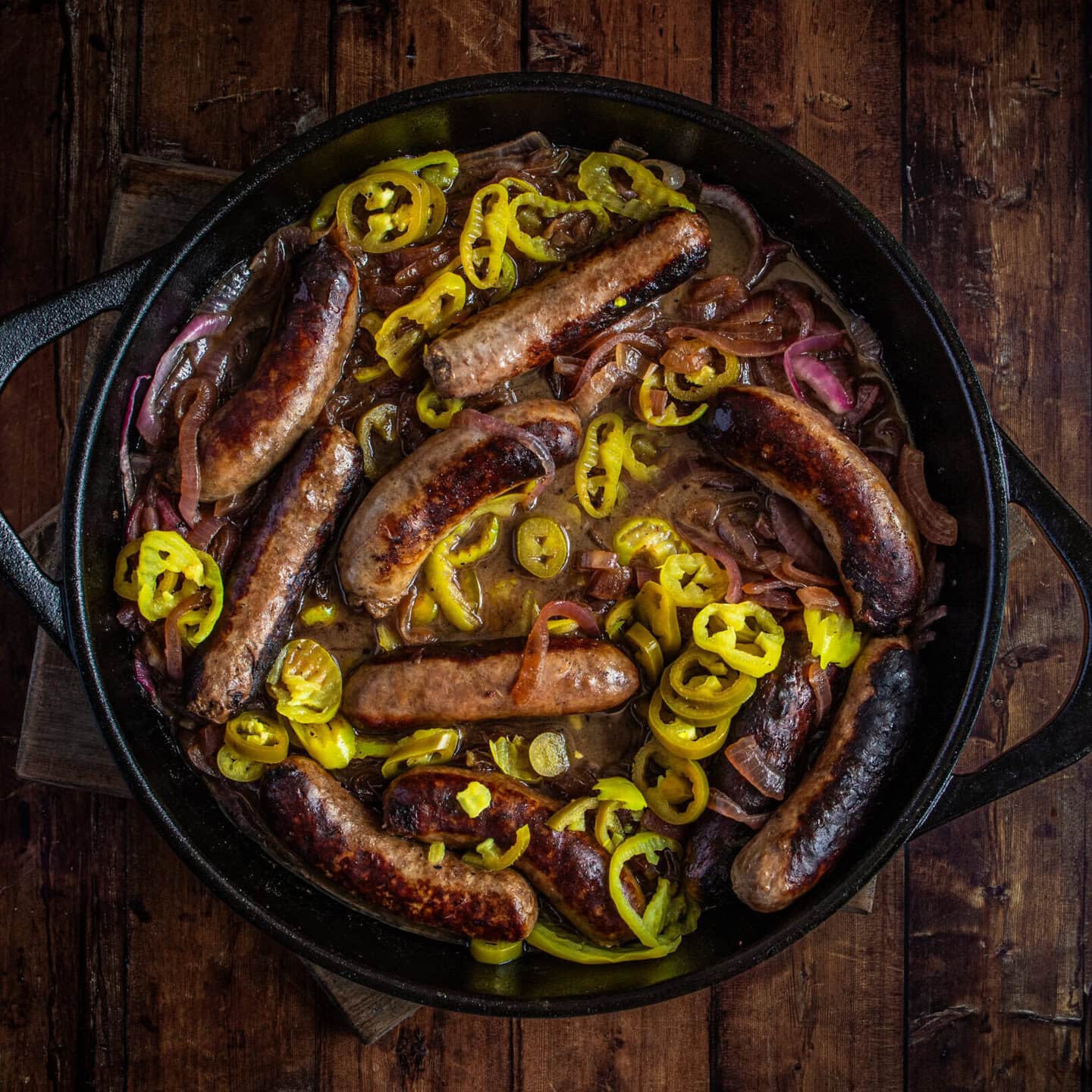 Fried sausages and peppers in cast iron black pan set on a wooden table.