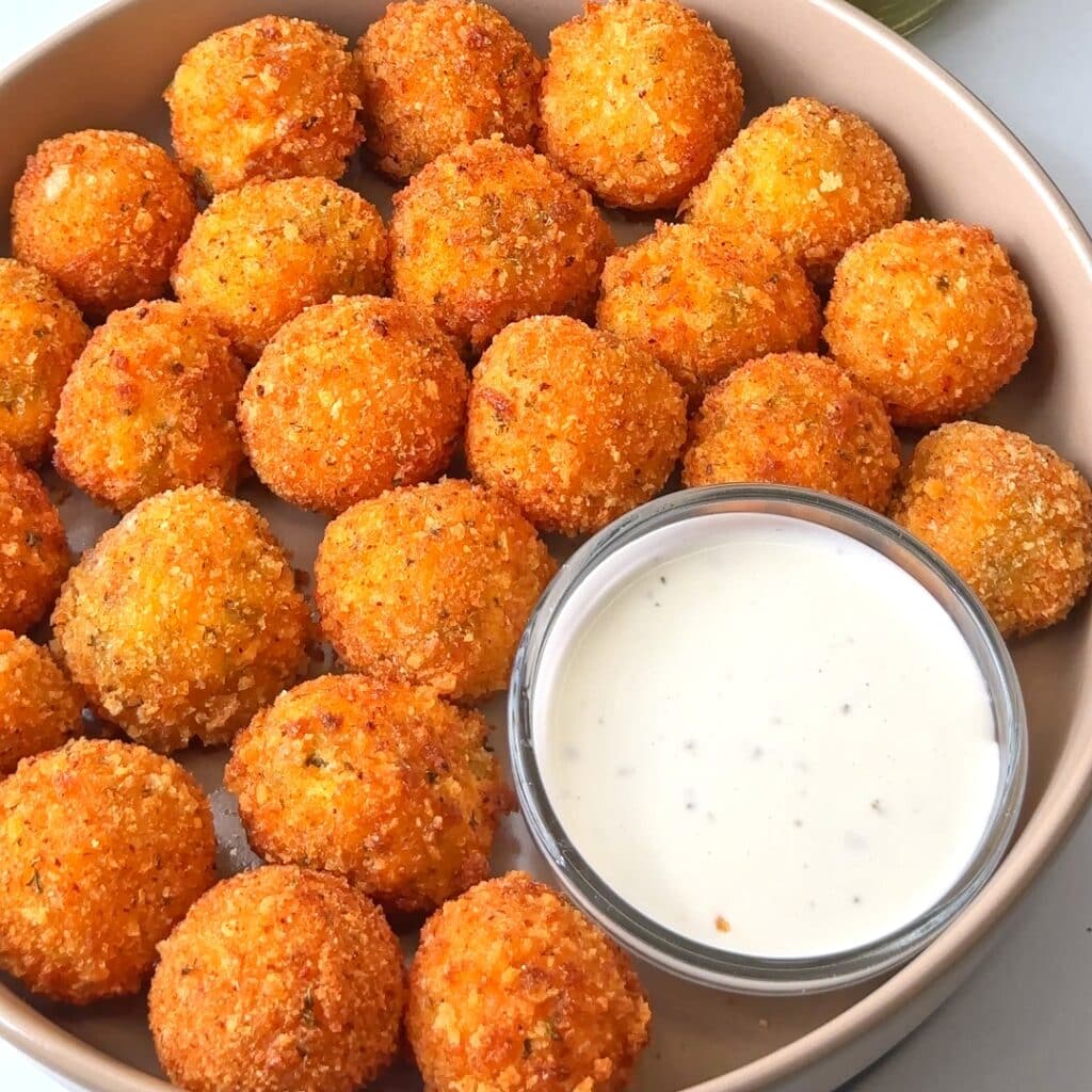 A bowl of crispy fried pickle cheeseballs with a side of dipping sauce.