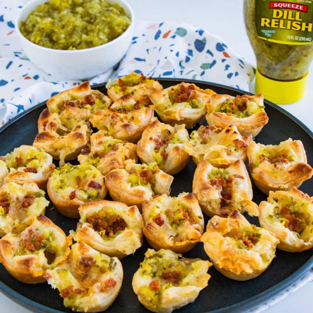 Pickle Brie Bites pastries on a black plate with Mt. Olive relish in background.