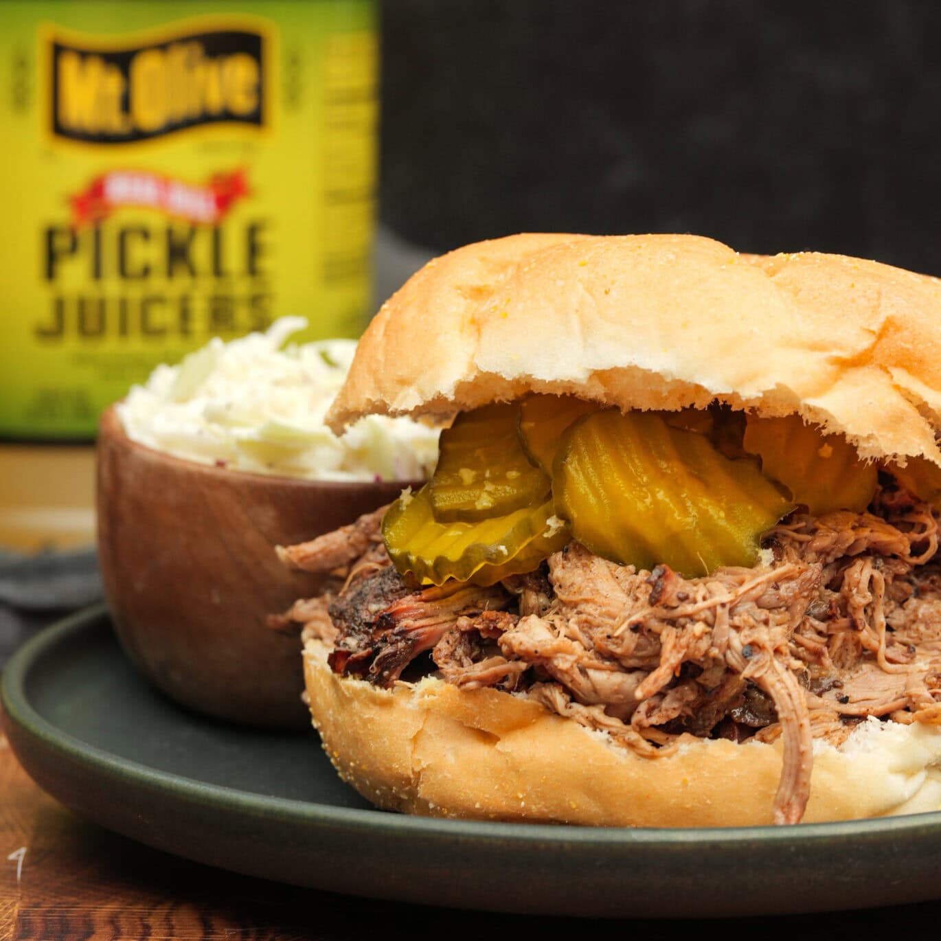 A plate with a Pickle Juicers Pulled Pork Sandwich topped with pickle slices and cole slaw.
