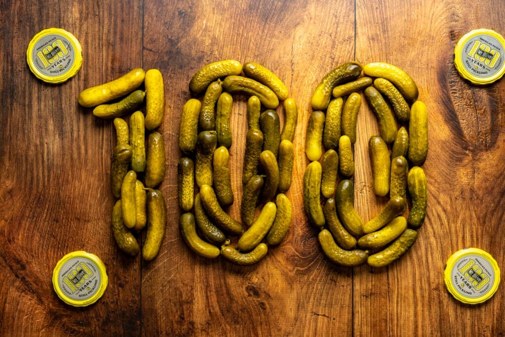 "100" spelled out in pickles surrounded by Mt. Olive Pickles Jar lids 100 Years Still Pickling