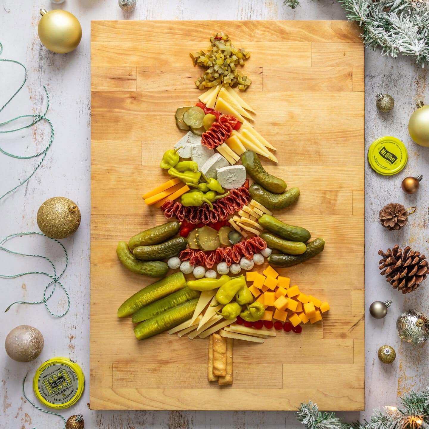 Wooden cutting board holding a Christmas tree-shaped arrangement of pickles, deli meats, and cheese cubes.
