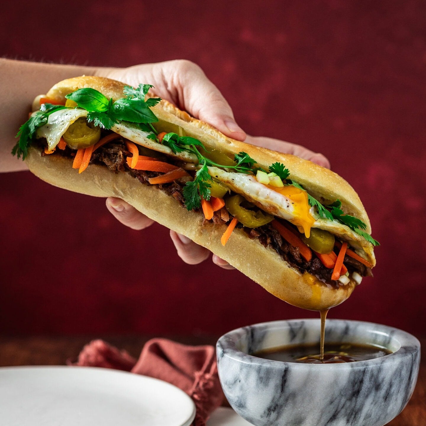 A hand dipping a French Dip Banh Mi sandwich into jus in a marble bowl.