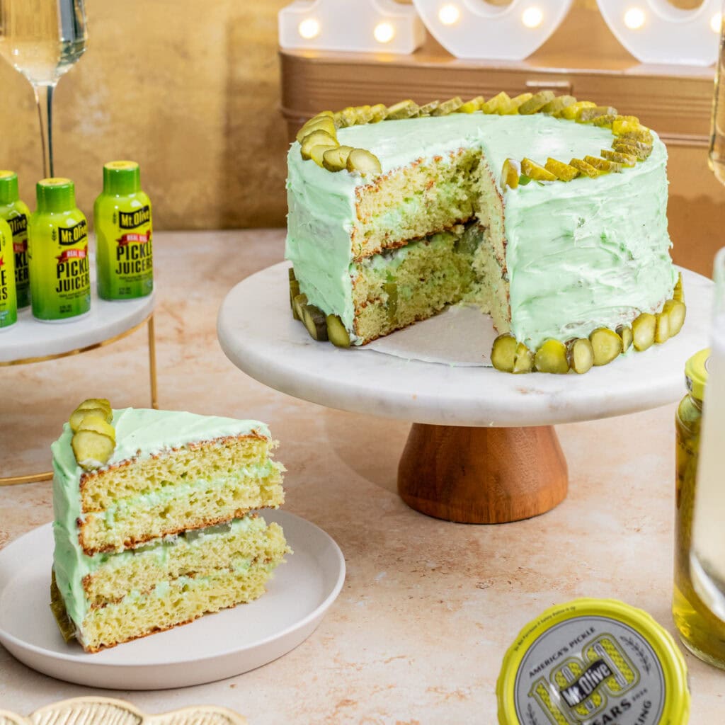 A pickle birthday cake on a cake stand with dill pickle juicers and separate slice of cake.