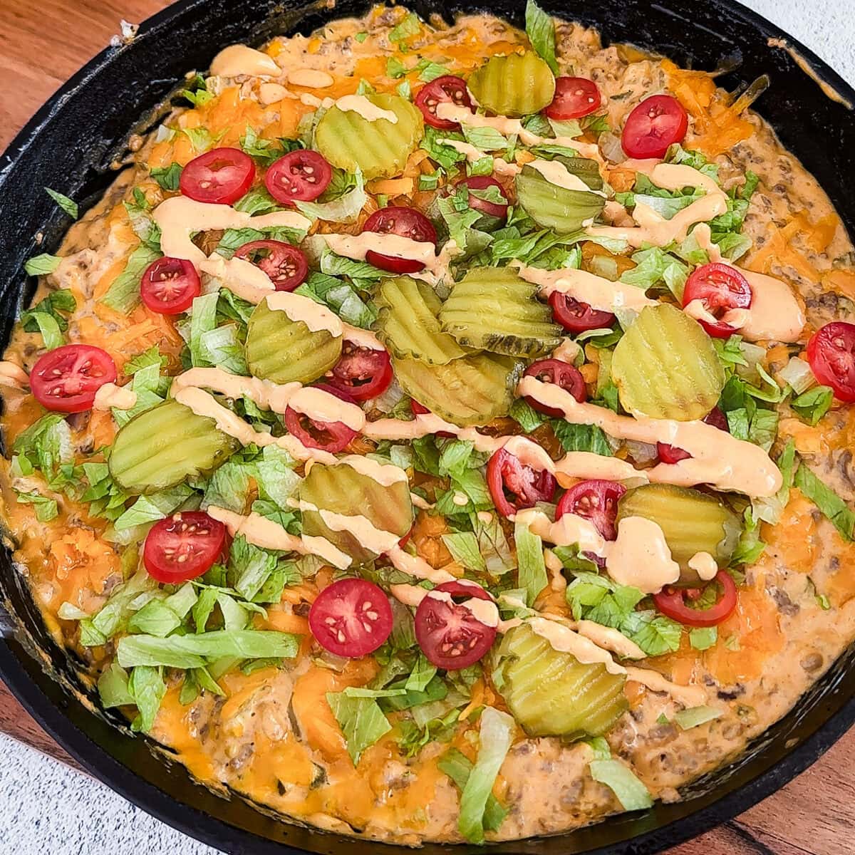 A skillet of Cheeseburger Dip topped with pickles, lettuce, and tomatoes.