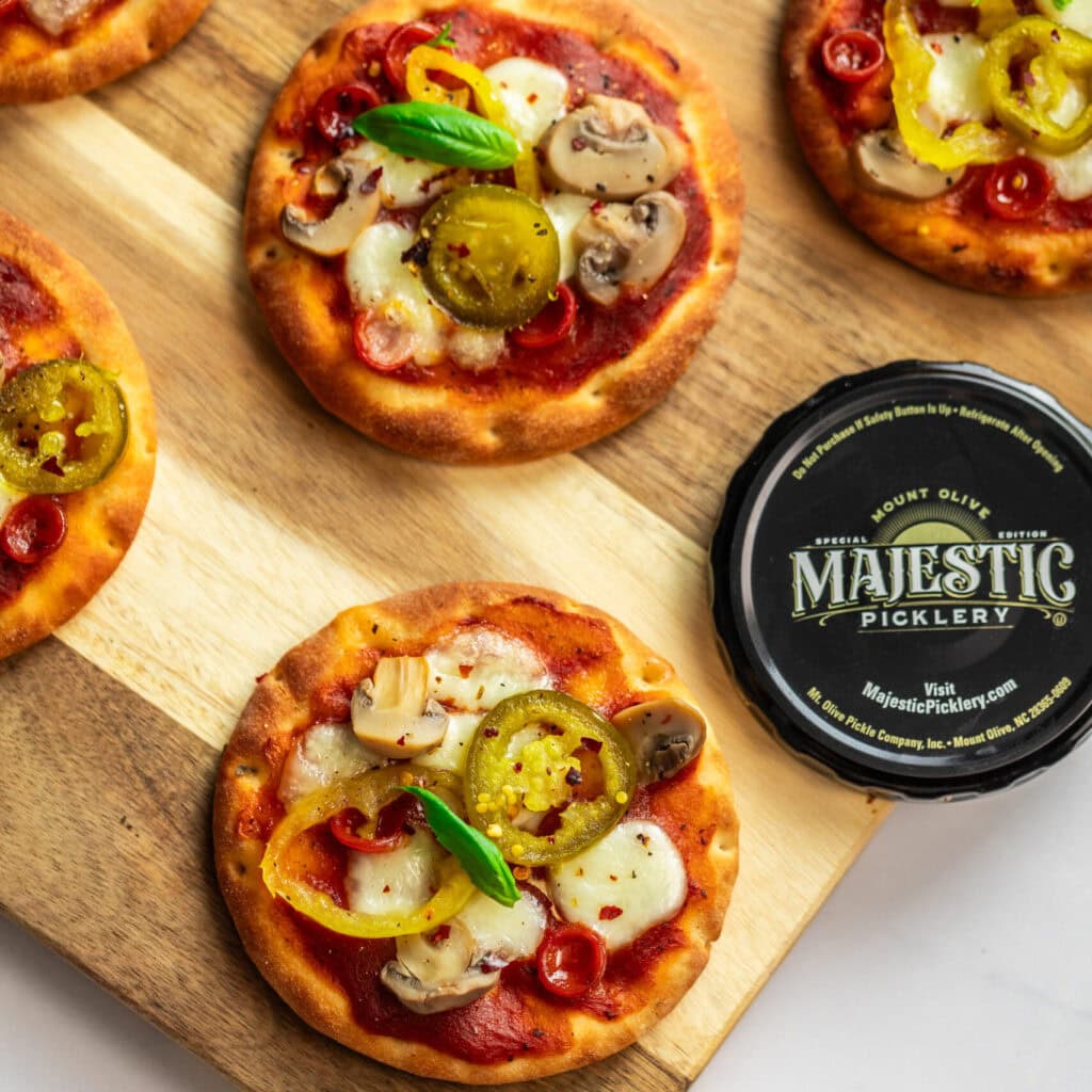 Mini jalapeno & mushroom-topped pizzas on a wood cutting board with Majestic Picklery lid.