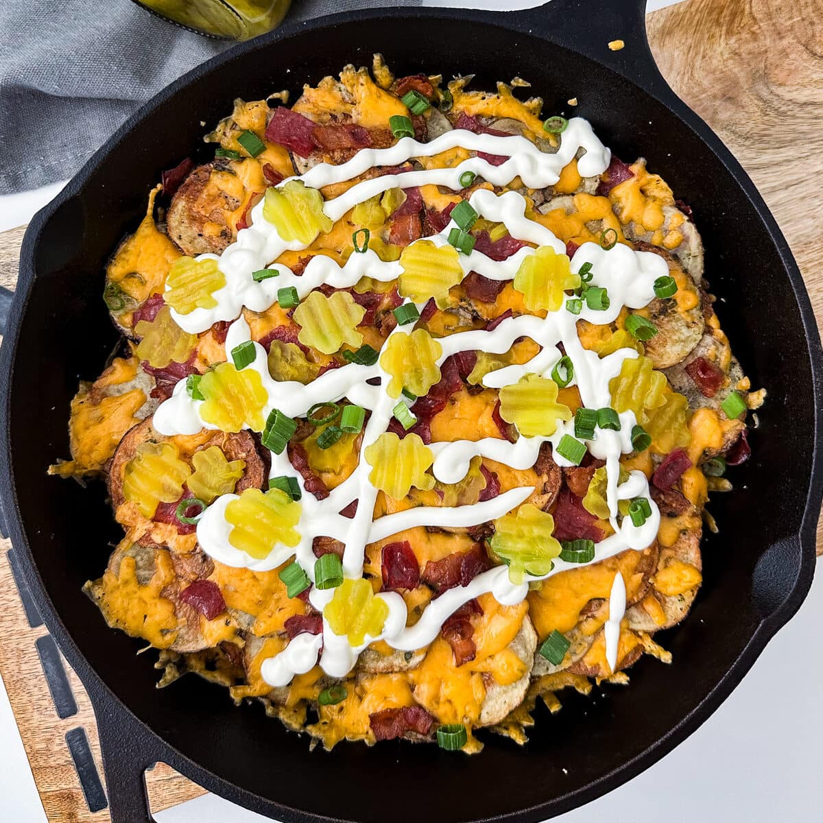 A cast-iron skillet with Irish potato nachos topped with shamrock pickles, bacon, and cheese.