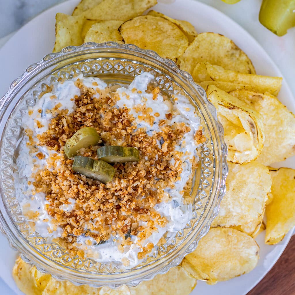 A glass bowl of fried pickle dip with crunchy potato chips on the side.