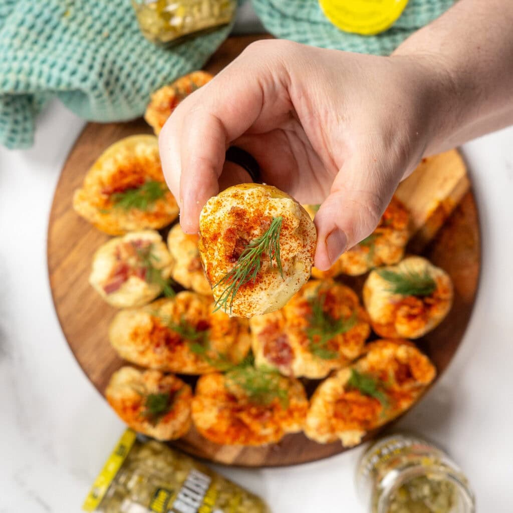 A hand holding a dill pickle deviled potato over a wooden plate of deviled potatoes.