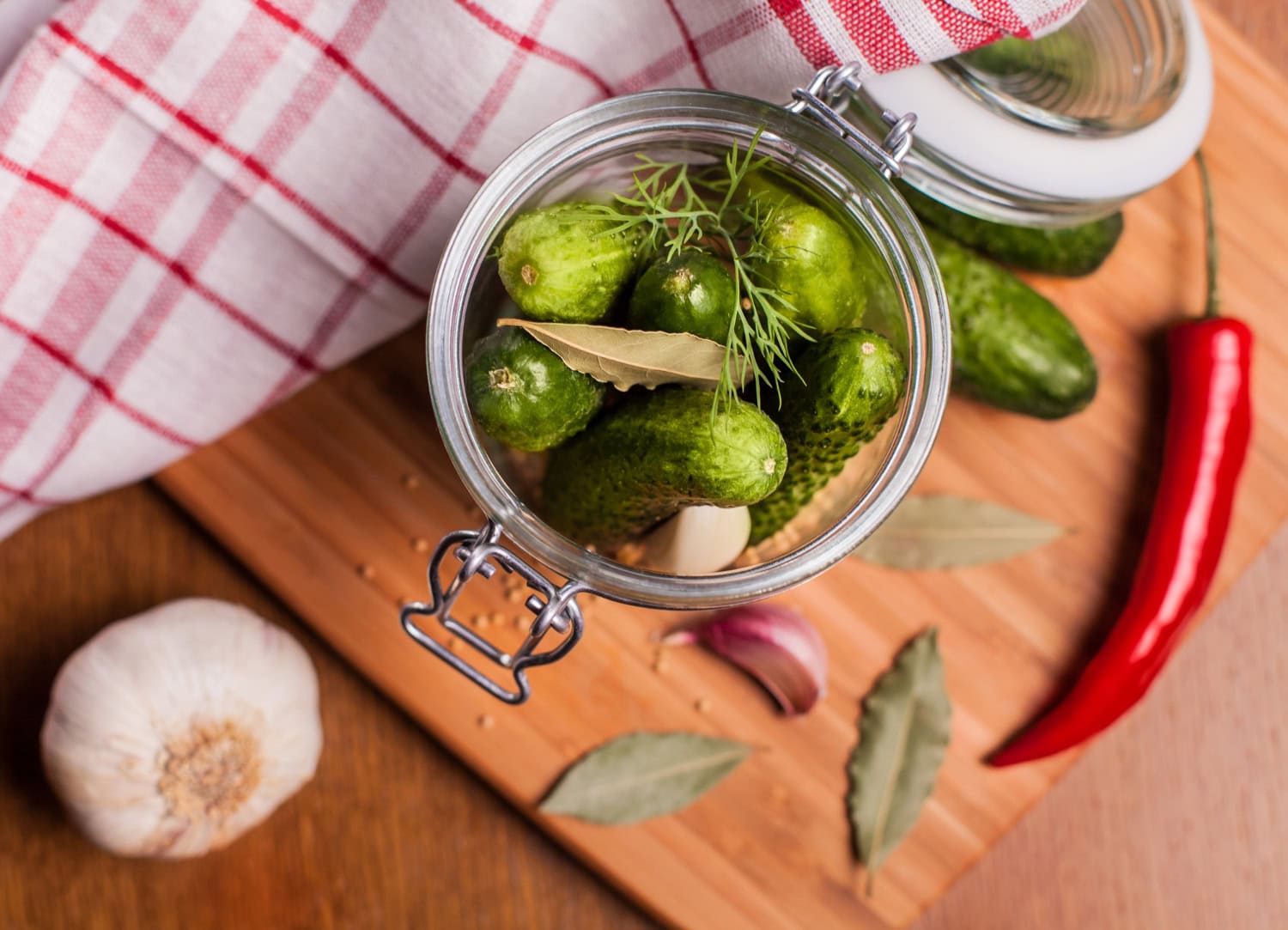 Jar of homemade pickles with cucumbers, garlic, and herbs on a wooden cutting board.