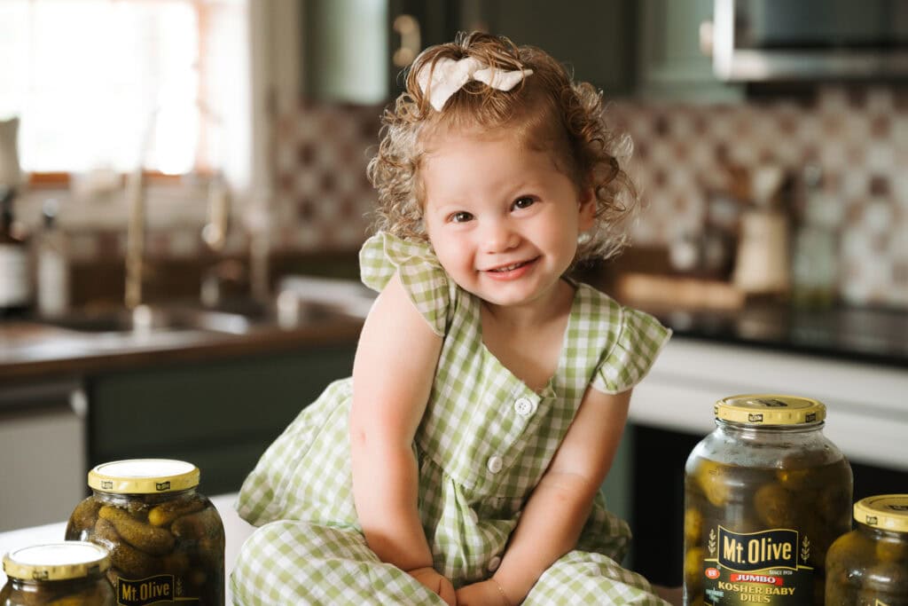 Cute girl with curly hair and green checkered dress in kitchen with Mt. Olive pickles.