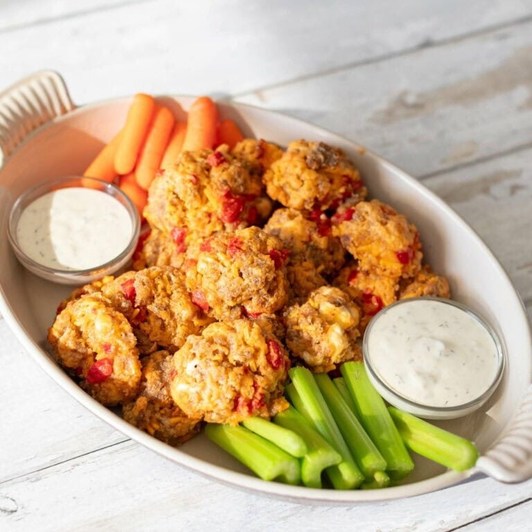 white table with large white platter dish. dish contains celery and carrots as garnish and two small dipping bowls of ranch dressing. middle of bowl contains sausage balls made of roasted red peppers and pimento cheese.