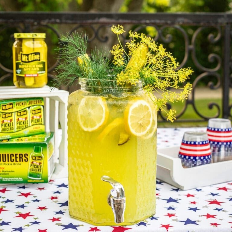 large glass beverage dispenser filled with lemonade and garnished with dill and pickles. 4th of july decor on table. Cases of Pickle Juicers