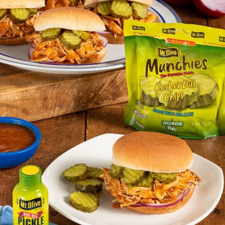 wood table. background shows multiple pulled chicken sliders on buns with pickles. Pouch of portable Munchies Pickles Kosher Dill Chips. White plate shows close up of pulled chicken sandwich topped with pickles. blue dish of bbq sauce. Pickle Juicer bottle in front.