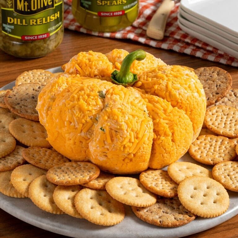 wood table. white place showing assorted crackers. middle of plate has a orange cheese ball shaped into a pumpkin. two jars of mt. olive pickles and relish in background.